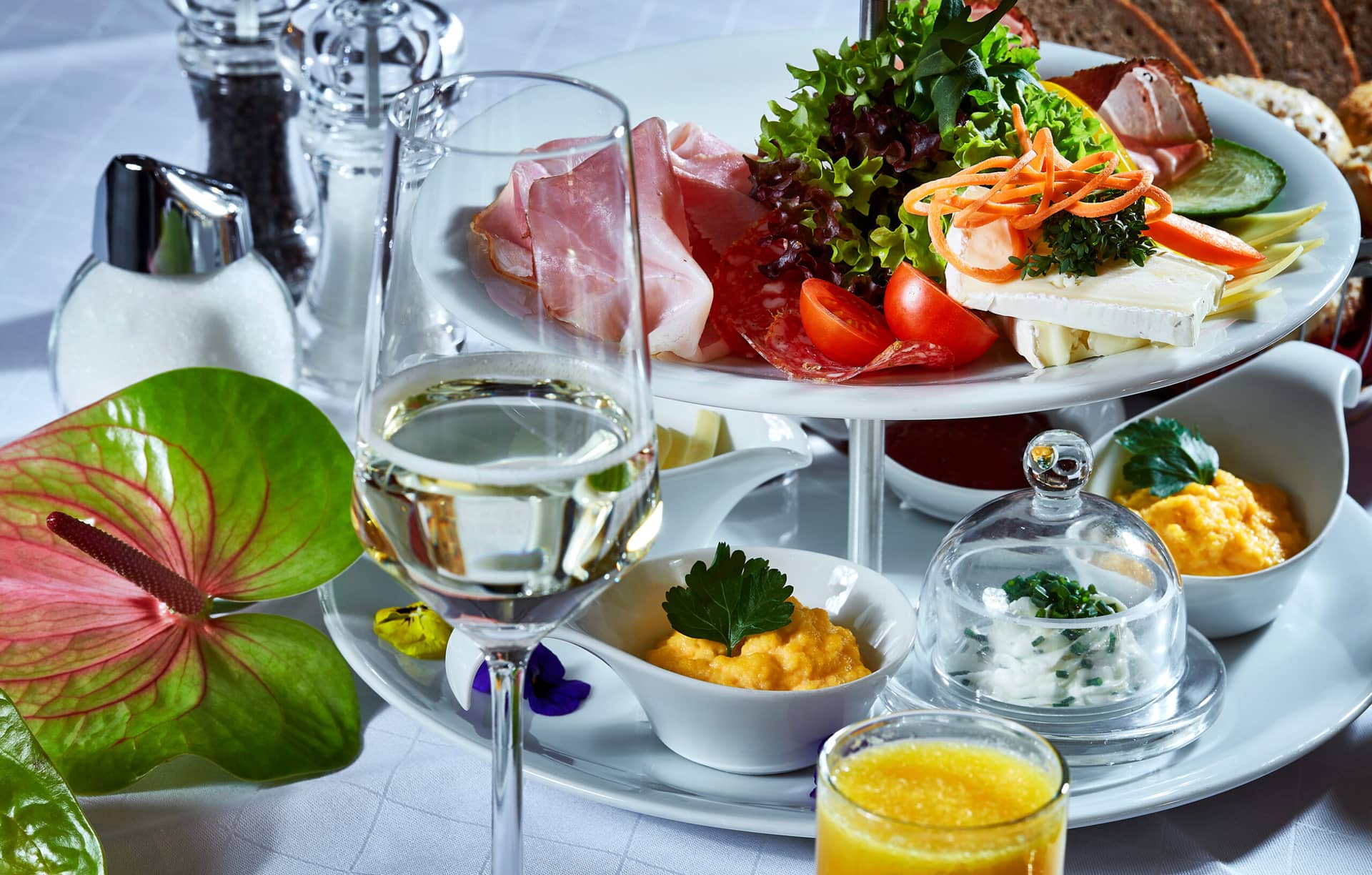 Ein gedeckter Tisch mit einer eleganten Frühstücksplatte im Traumwerk, mit Aufschnitt, Käse, frischem Gemüse, Rührei, Aufstrichen, einem Glas Weißwein, Orangensaft und verschiedenen Gewürzen auf einer weißen Tischdecke - perfekt vor Ihrem Porsche-Ausflugsziel.
