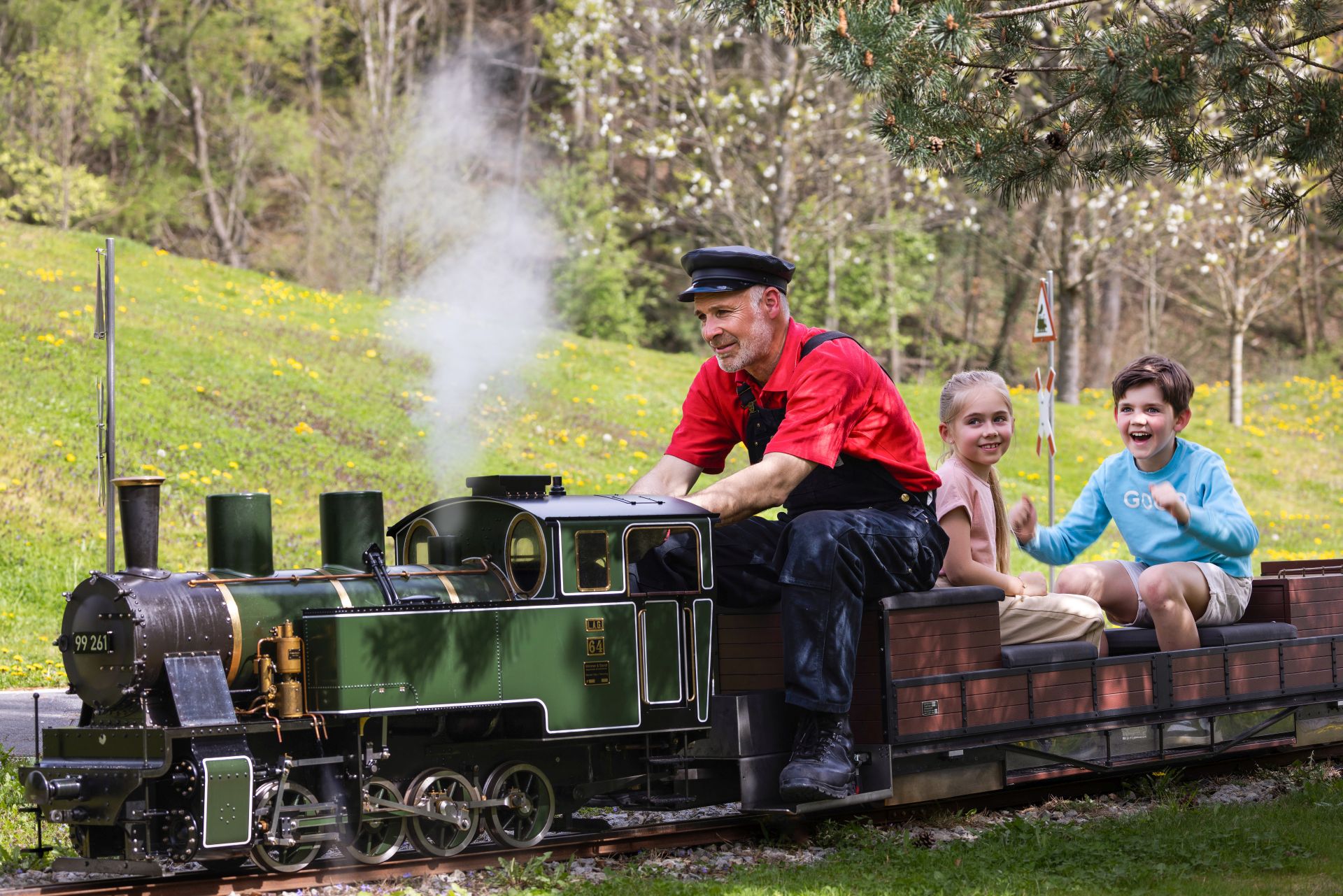 Ein Mann in Schaffneruniform fährt eine kleine Modelleisenbahn im Freien, hinter ihm zwei lächelnde Kinder an einem sonnigen Tag in einem grasbewachsenen, bewaldeten Park - ein ideales Ausflugsziel für Familien.