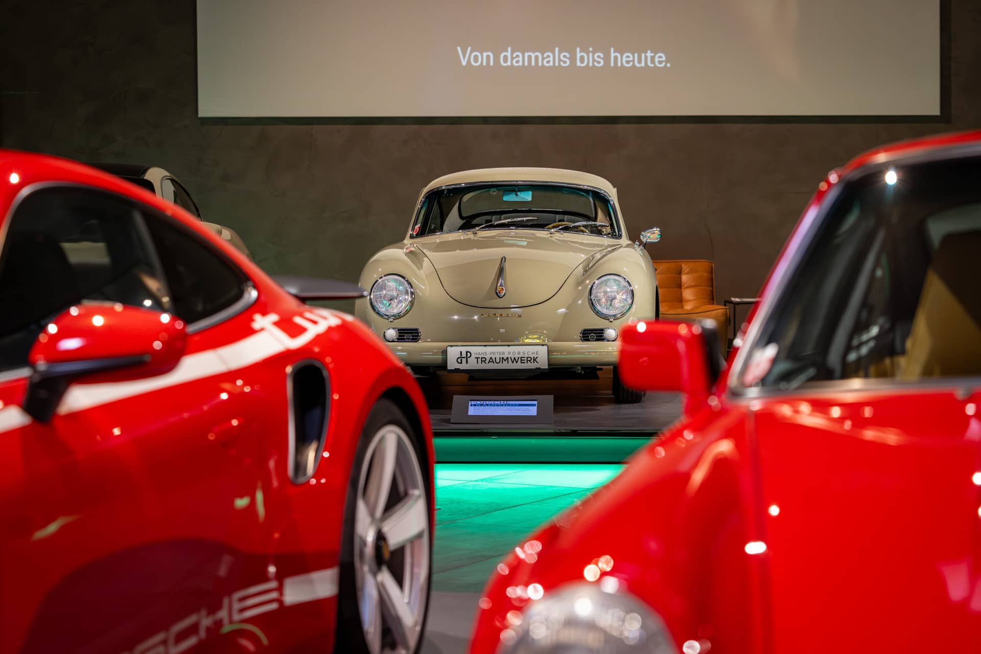 Ein klassischer cremefarbener Porsche steht in einem Ausstellungsraum auf einem Podest zwischen zwei modernen roten Porsche-Fahrzeugen, darüber wird der deutsche Text Von damals bis heute projiziert.